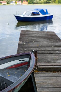 Weathered rowboat moored beside a wooden dock, with a blue motorboat softly blurred on the calm lake in the background.