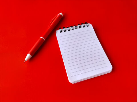 Small white spiral notebook with lined pages placed next to a matching red ballpoint pen.
