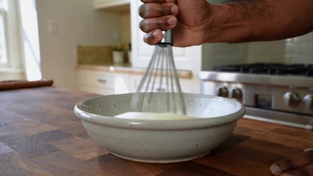 Whisking cook's hands starting upright to aerate batter with steel whisk in bowl in home kitchen
