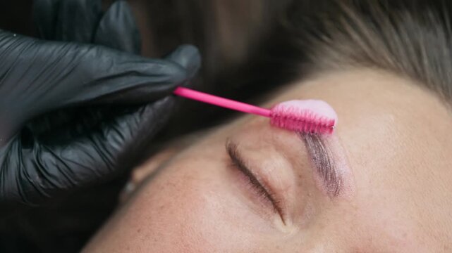 Close-up of a beauty specialist applying lamination solution to a clients eyebrow with a spoolie brush during a professional brow treatment.