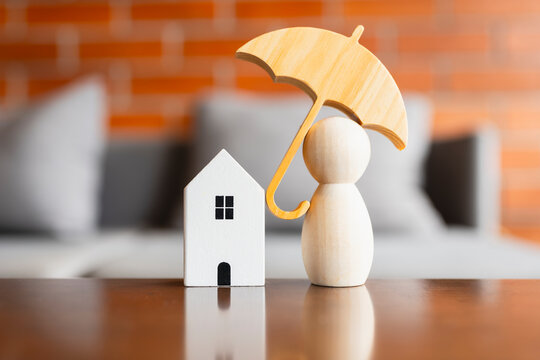 Home and life insurance concept with wooden umbrella covering miniature house model and peg doll figure on reflective table with brick background for property mortgage safety and protection.