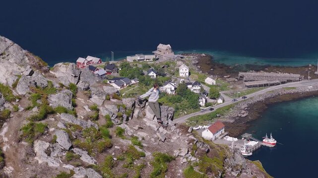 Aerial view of a man resting on a rocky mountain ridge overlooking a coastal village and clear turquoise sea in Lofoten, Norway.