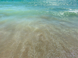 Waves break gently on a sandy beach under a bright sky, creating a scene of sun and water during midday © DSMT