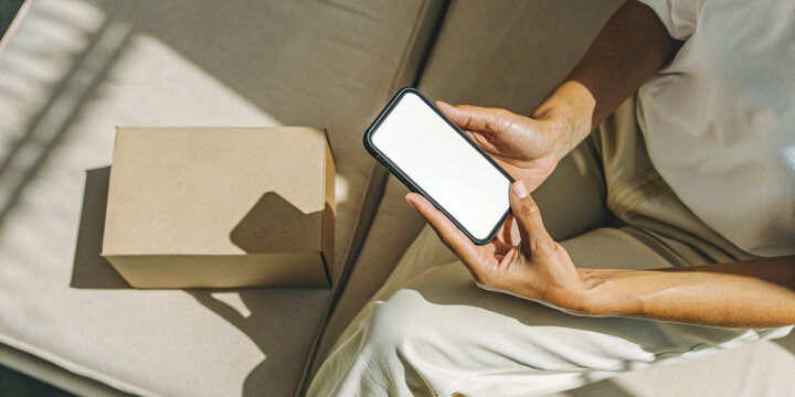 Online shopping on a mobile app with someone holding a smartphone with a white screen next to a cardboard box on the sofa