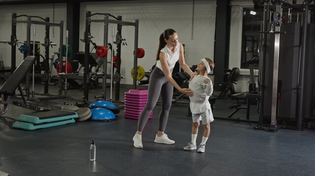 Wide shot of Caucasian young adult woman training Caucasian male child in gym, demonstrating shoulder stretches. Clip is ideal for family fitness and healthy lifestyle campaigns