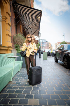 Stylish mature woman with suitcase using smartphone on city street