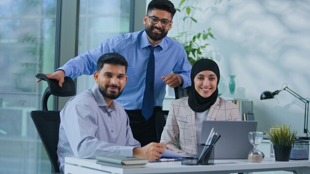 Muslim business team three Arabian businesspeople smiling at table satisfied with working good results with computer in office Indian co-workers work online smile looking at camera together in agency