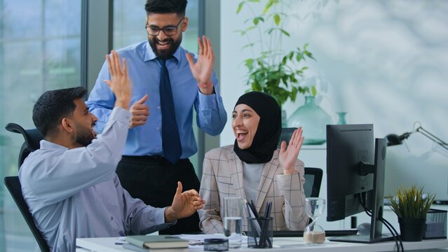 Arabian business team with papers and computer celebrating success giving high five happy with goal in office Indian men and Muslim woman looking at computer checking document celebrate startup growth