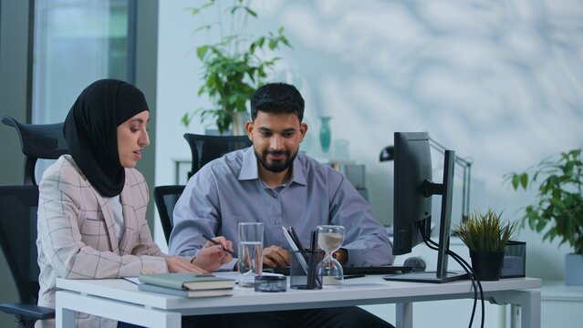 Two Indian business people team in office working with computer and paperwork smiling talking planning project strategy Arabian colleagues talk work with computer woman in hijab teamwork with man
