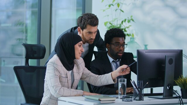 Multiethnic business team three colleagues discussing talking looking in computer together in office Caucasian leader boss help advise talk helping Muslim woman and African American man with work