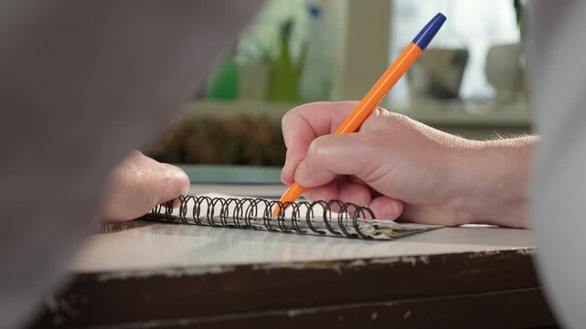 Woman's hand writes in notebook with ballpoint pen while sitting at desk in office doing paperwork