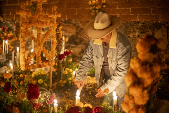 Patzcuaro, Michoacan, Mexico - 01 November 2023: View of a man in a cowboy hat lighting candles at a grave adorned with marigolds during the Day of the Dead celebration.