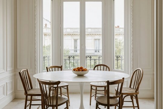 Parisian dining room featuring a round white marble table, vintage wooden chairs, and a bowl of clementines by large bay windows