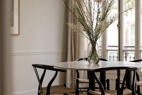 View through a blurred black wishbone chair towards a round marble table adorned with a vase of dried flowers in a Parisian dining room