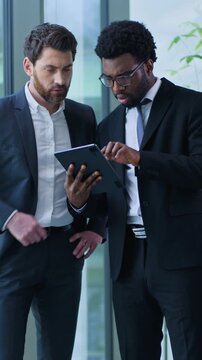 Two multiethnic business men talking looking in digital tablet device discussing work startup online project in office multiracial businessmen males agents talk working together with wireless gadget