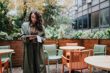 A businesswoman stands outside a cafe reading a tablet while holding a sheet of paper. She reviews documents in a quiet outdoor seating area surrounded by chairs and planters. © qunica.com