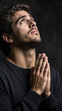 Faithful young man praying with hands together and looking up. Spiritual devotion and religious practice in low light setting.