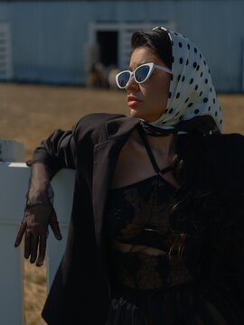 Asian woman in jacket, headscarf and sunglasses posing near farm fence