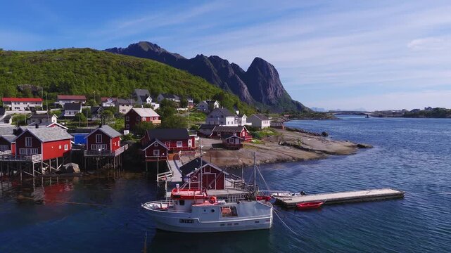 Aerial view of traditional red rorbuer cabins and a fishing boat moored at a dock in Reine, Lofoten Islands, Norway, on a sunny day.