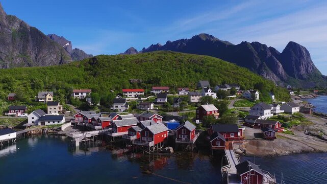 Aerial view of traditional rorbuer cabins and village houses along the fjord coast with mountains in Reine, Lofoten, Norway on a clear day.