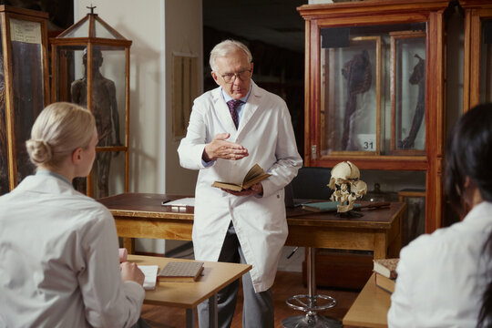 Senior Caucasian male professor holding book and lecturing young adult students in anatomy museum. Image suitable for medical education and university recruitment campaigns