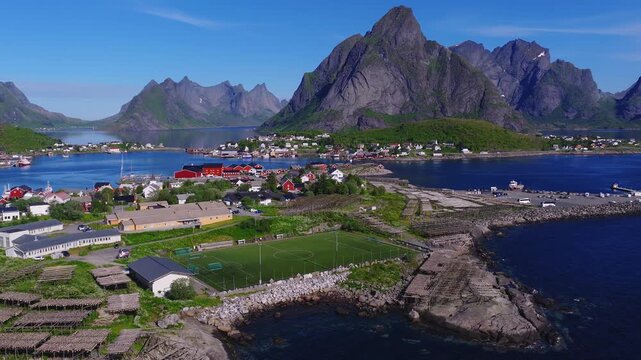 Aerial video showing the coastal fishing village of Reine in Norway, surrounded by sea water, rocky mountains, and traditional drying racks on a sunny day.