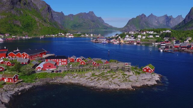 Aerial view gliding over traditional red rorbu cabins, drying racks, and rocky coastline surrounded by fjords and mountains in Reine, Lofoten Islands, Norway.