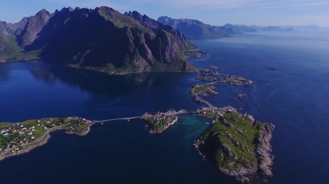 Aerial view shows the bridges connecting small islands and fishing villages with steep mountain peaks in Lofoten, Norway on a clear sunny day.