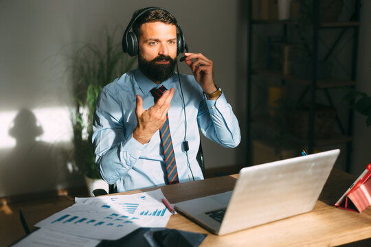 Bearded businessman wearing a headset with a microphone, gesturing while participating in an online video call from his home office, utilizing a laptop for remote work and telecommunication