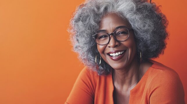 Smiling African American woman with curly gray hair wearing glasses, dressed in orange top, against vibrant orange background, exuding warmth and confidence in a joyful portrait