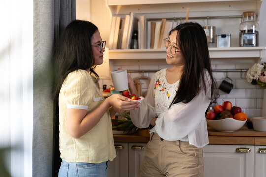 Two young women share coffee and strawberries while chatting in a cozy kitchen corner. The candid moment represents friendship, comfort, and healthy everyday living