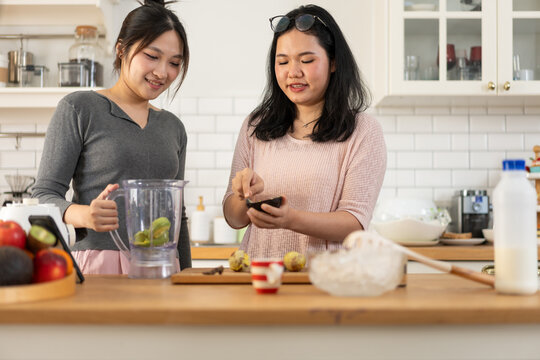 Two women prepare a fresh fruit smoothie together while using a smartphone for recipe guidance. The lifestyle scene blends healthy eating, technology, and everyday home cooking