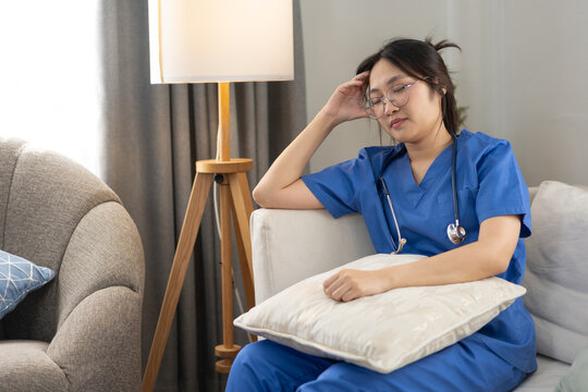 A female healthcare worker in blue scrubs rests on a sofa while holding a pillow after a demanding workday. The lifestyle portrait represents nurse burnout, exhaustion, and the importance of self-care