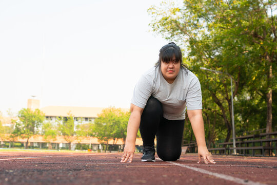 Plus size woman preparing to sprint on an outdoor running track with focused posture, promoting perseverance, fitness goals and healthy living in a bright park setting with generous copy spa