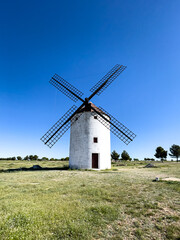Fototapeta premium A centered shot of a single traditional white windmill with large wooden lattice sails standing on a grassy meadow under a clear blue sky.