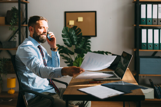 Confident bearded businessman busy working in his office, talking on a smartphone and organizing documents, creating a dynamic scene of multitasking and productivity