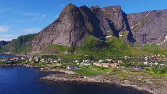 An aerial view reveals the coastal football field, traditional buildings, and massive mountain cliffs in Reine, Lofoten Islands, Norway.
