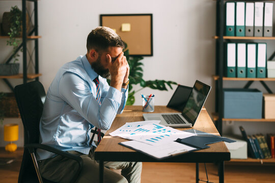 Businessman feeling overwhelmed and experiencing stress while working with documents and a laptop in a modern office, symbolizing professional challenges and mental exhaustion