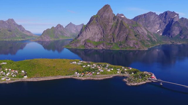 An aerial view over calm fjord waters, coastal village houses, and steep mountain peaks in Lofoten, Norway during clear daytime.