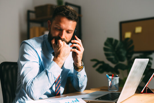 Bearded man sitting at his desk, having a tense phone conversation while biting his nails, reflecting stress, anxiety, and deep thought in a professional office environment