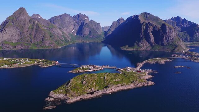 An aerial panorama reveals the coastal fishing village of Reine, surrounded by calm fjord waters, bridges, and rocky mountains on the Lofoten islands in Norway.