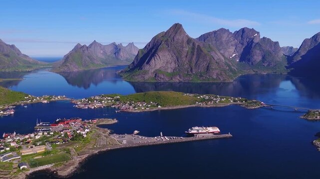 An aerial view reveals a passenger ship moored at the dock in Reine, surrounded by calm fjord waters, coastal bridges, and mountains in Lofoten, Norway.