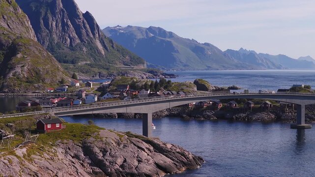 Aerial view of a coastal road bridge connecting rocky islands and traditional rorbu cabins in Reine, Lofoten, Norway on a clear day.