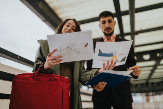 Two business colleagues review printed financial charts and a timeline slide while standing in a modern transit hub. They examine graphs and documents together, discussing data and project progress.
