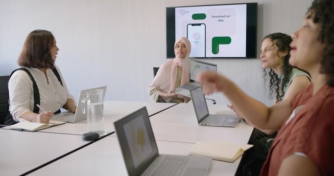 Female coworkers listening as presenter in scarf introducing app on laptop monitor seeking feedback
