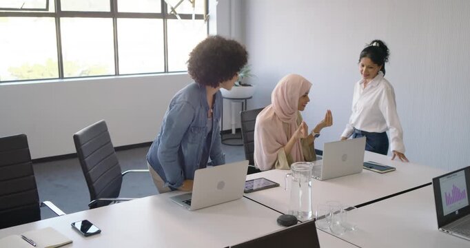 Diverse female team gathering around hijab woman after presenting laptop chart, clapping in office