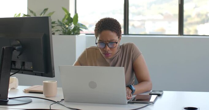 Woman sitting at desk in bright office, glancing down, placing hands, typing report on laptop