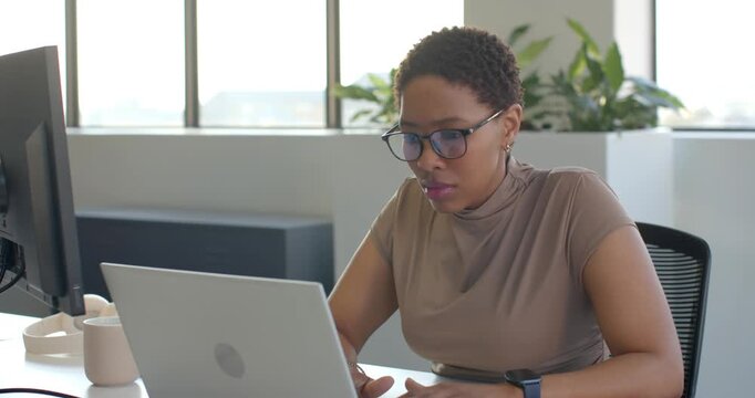 African woman typing, glancing at laptop and external monitor, pointing after spotting detail