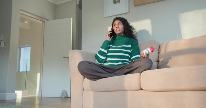 African American woman on phone, checking white bottle red cap to confirm dosage on sofa