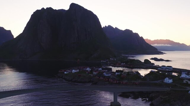 Aerial view of the fishing village of Reine in Lofoten, Norway, showing coastal bridge, rorbuer cabins, and steep mountain peaks at dusk.